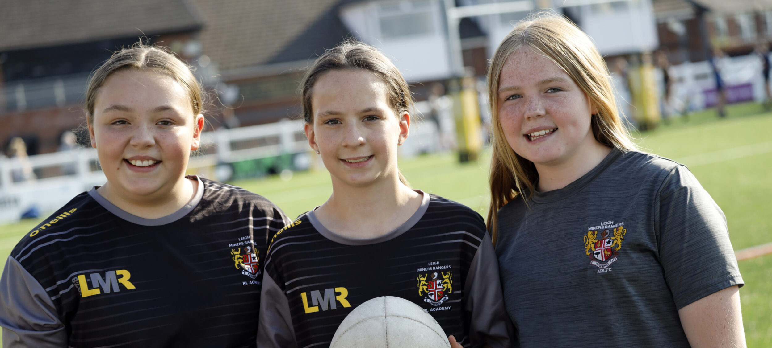 Three girls holding rugby ball and smiling at Leigh Miners Rangers Rugby Club